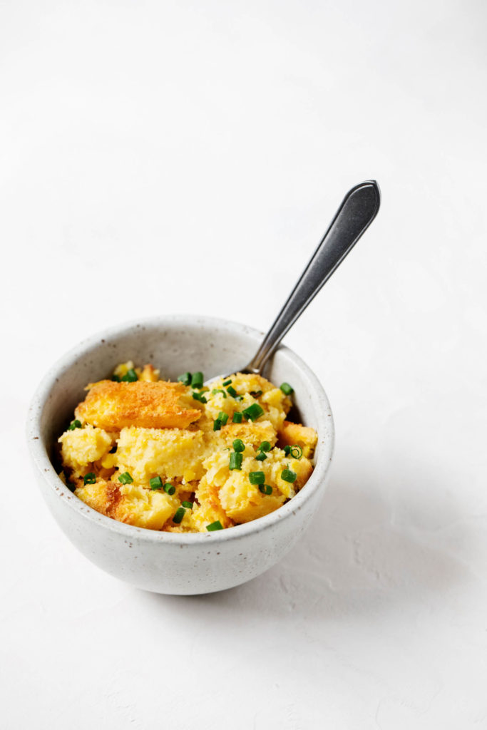 A small bowl of soft, vegan corn pudding. Ready to eat, with a serving spoon