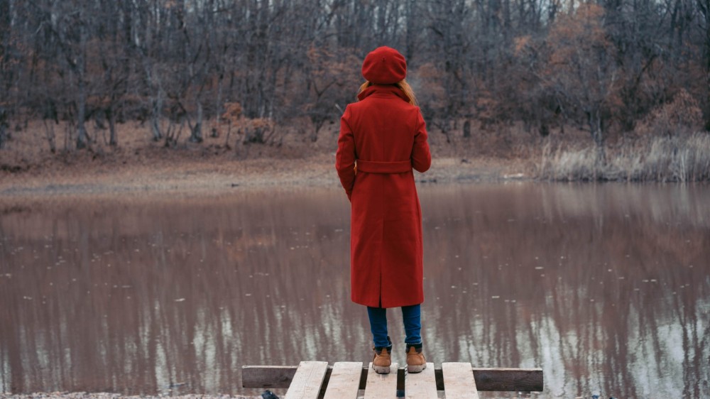 A woman in a red trench coat stands on a dock and looks out at a pond in cold weather.