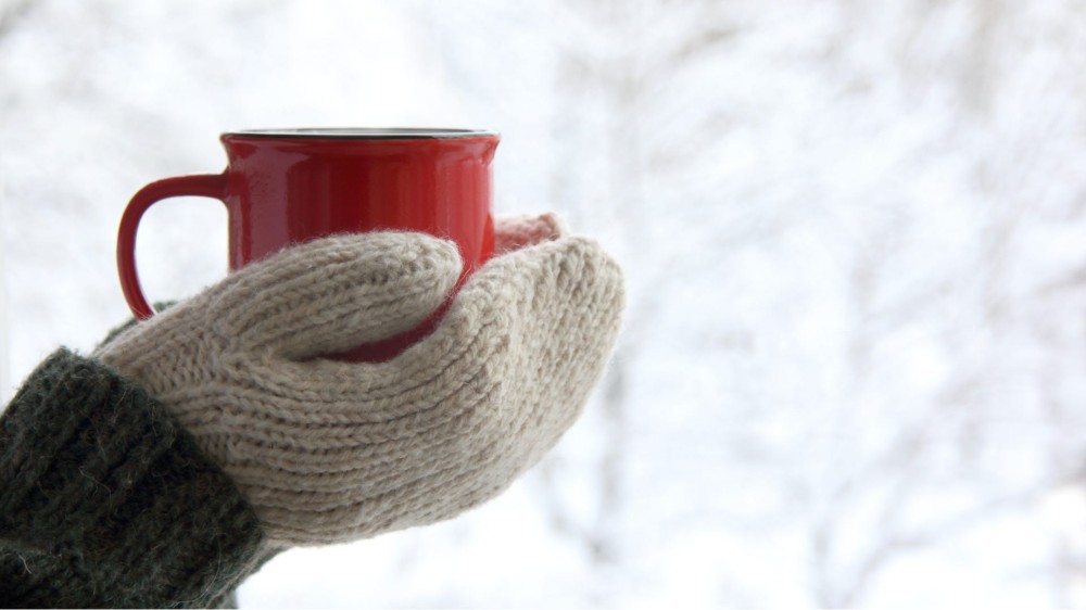 A man holds a mug while wearing mittens out in the snow.