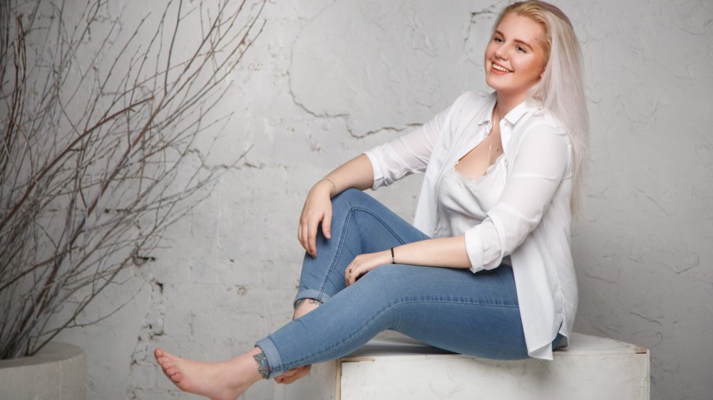 A blonde woman wearing jeggings and a white shirt sits on a wooden platform.