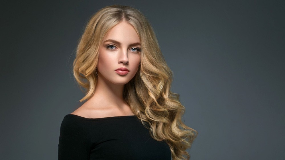 A woman with an oval face shape, and long, wavy blonde hair with a center part. 