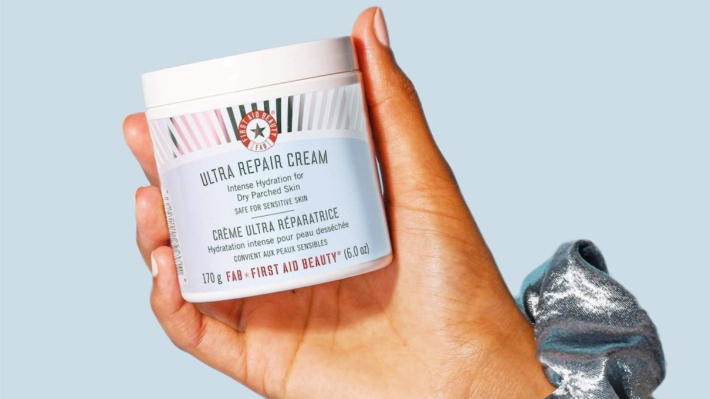 A hand holding a container of First Aid Beauty face cream. 