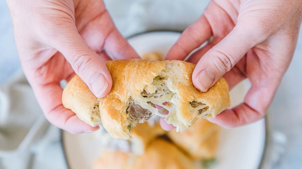 Someone holding a cheesesteak croisant and pulling it apart slowly to display the cheesy deliciousness in each bite.