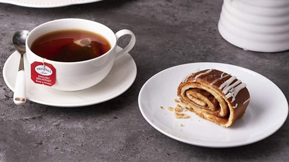 A cup of Twinings tea next to a cinnamon roll on a plate.