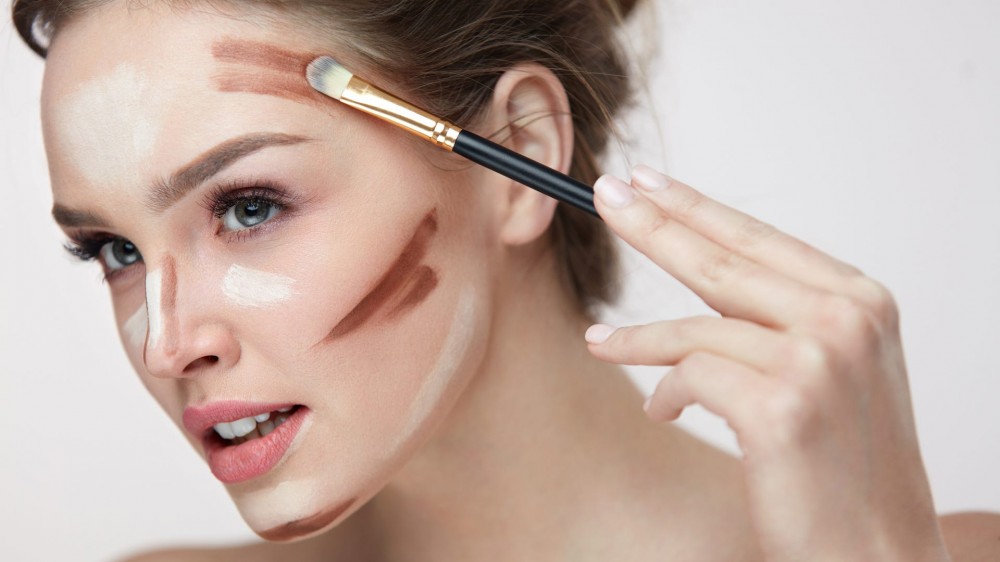 a young woman applies darker contouring makeup to her temple, cheekbone, chin, and side of nose and highlight to her forehead, under-eyes, and jaw.