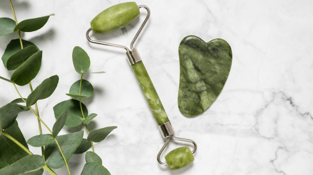 A green jade roller and gua sha stone for facial massage and eucalyptus branch on marble background.