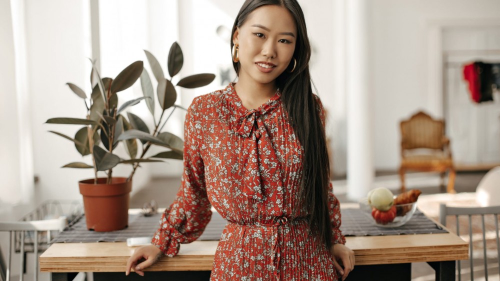 Long-haired brunette Asian woman in floral red dress smiles widely, leans on wooden table at kitchen and looks into camera.