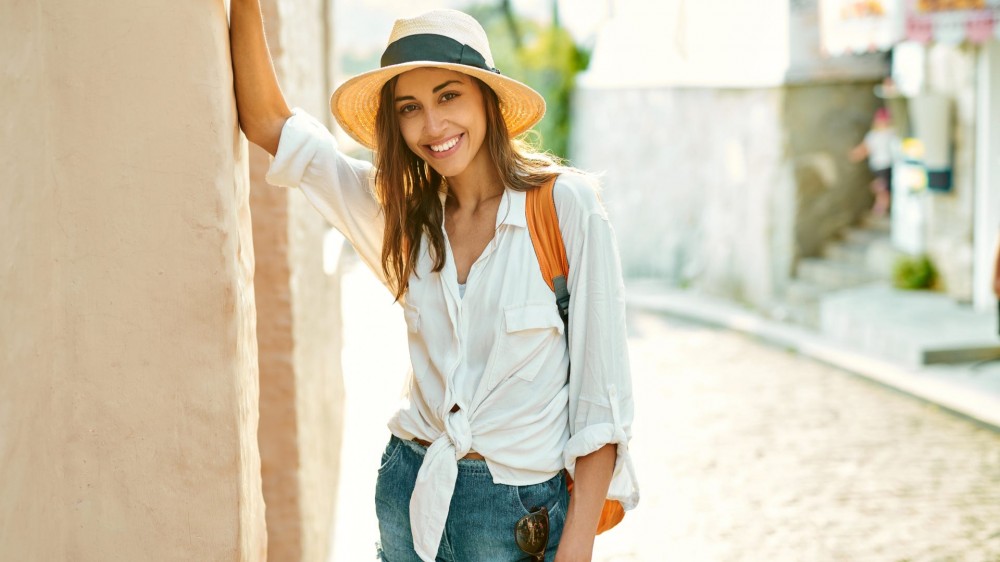 A cheerful tourist woman wearing a straw hat stands by a wall in an old town.