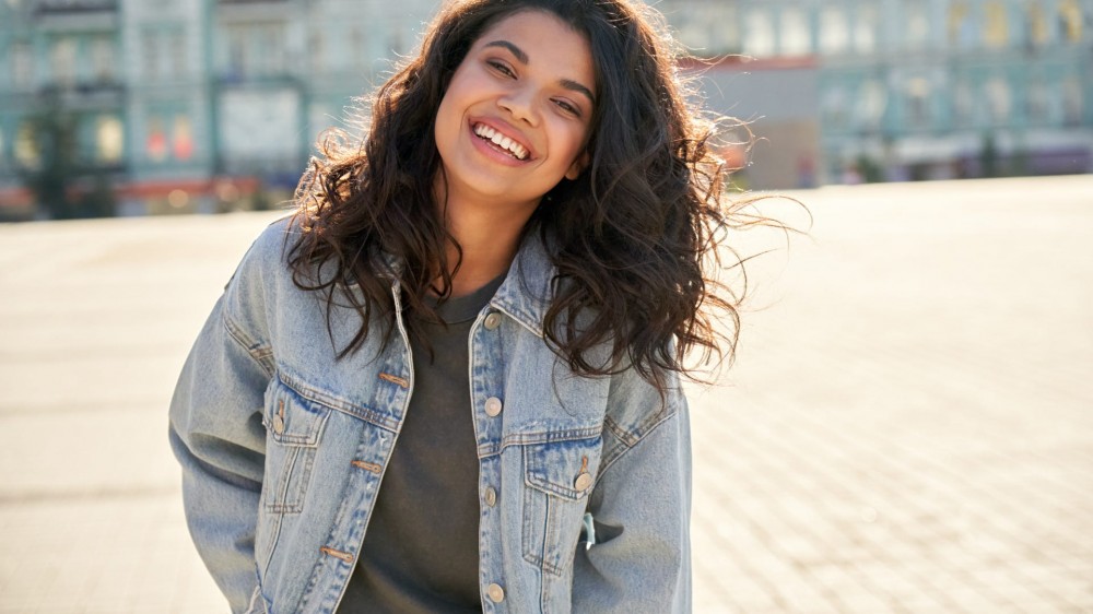 Happy African young woman wearing denim jacket laughing looking at camera standing on street.