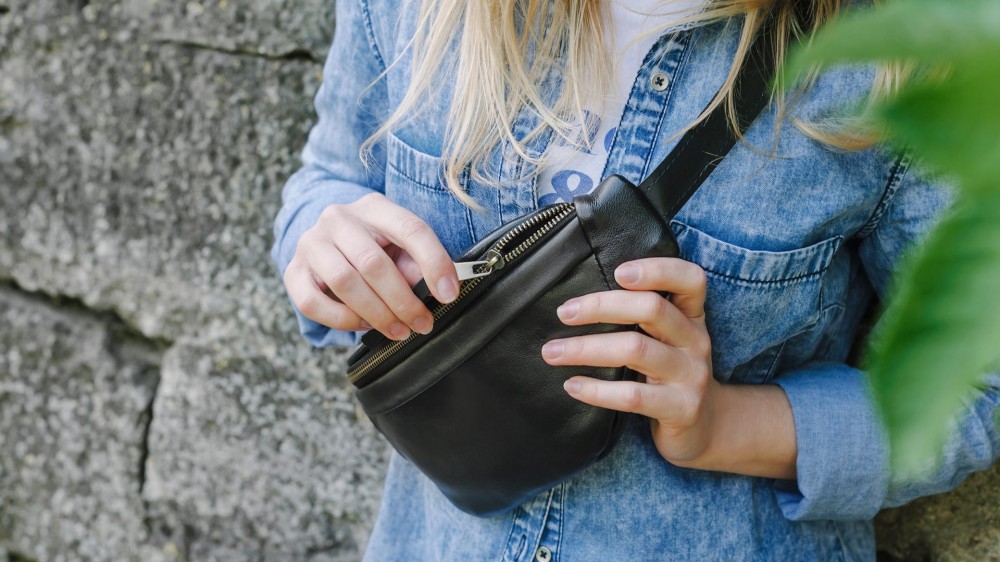 Blonde girl in a blue denim shirt is holding a black crossbody fanny pack.