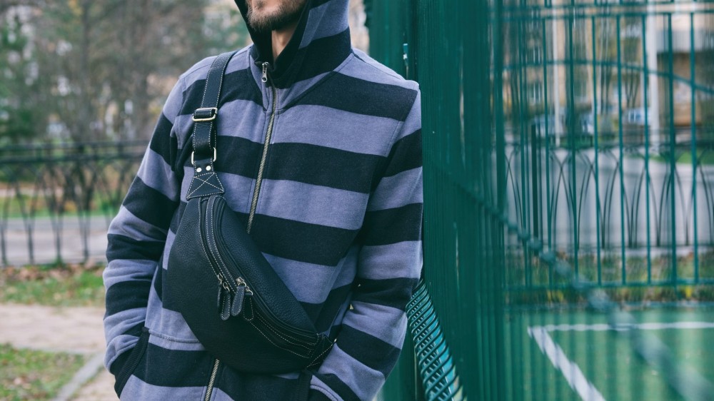 Cropped portrait of young bearded man in striped hoodie with black leather crossbody bag.