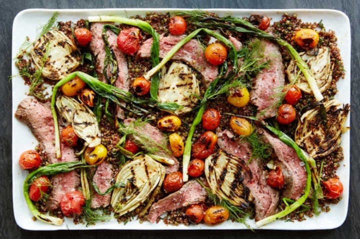 Grilled and sliced steak cooked to a perfect medium-rare, arranged on a rectangular plate with grilled fennel bulbs, cherry tomatoes and spring onions on on top of a bed of quinoa.