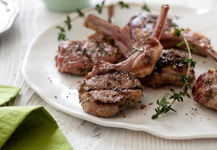 A white plate with a dainty pile of grilled lamb chops and garnished with fresh thyme.