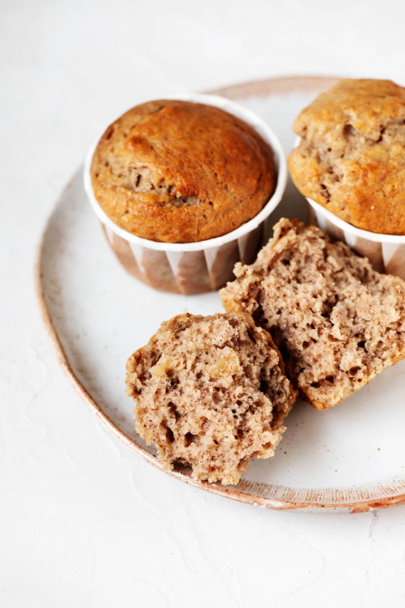 A small dessert plate holds vegan banana walnut muffins. Two are intact, in their liners, while a third has been broken in half.