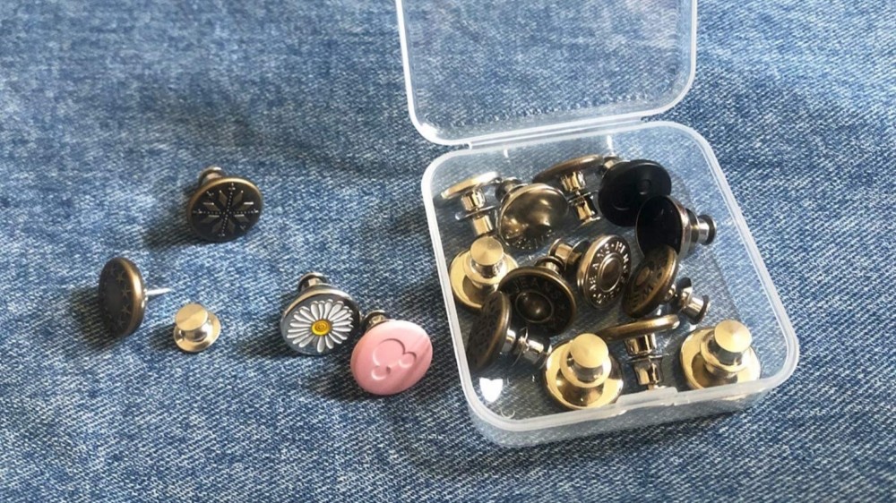 A clear opened button box with many button pins inside and four button pins sitting next to it. 