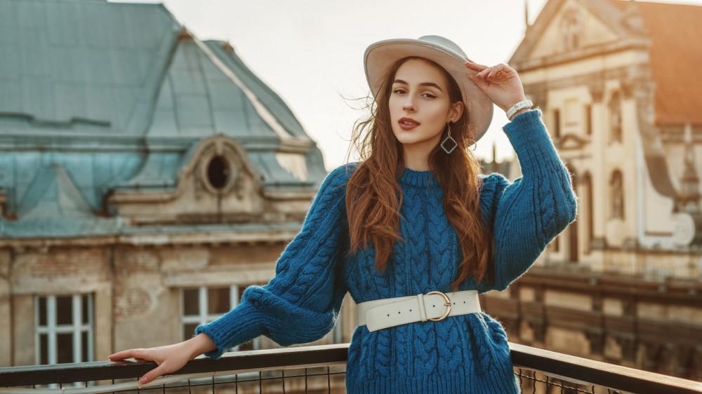 A woman wearing a blue sweater dress, a white belt around her waist, and a white hat.