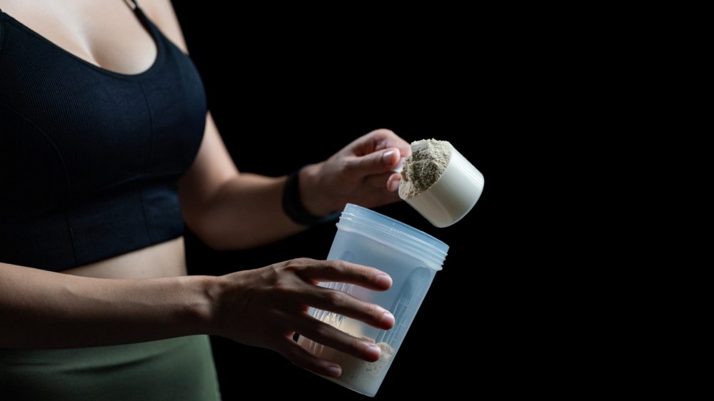 A young woman putting a scoop of protein powder into a shaker bottle after a workout.