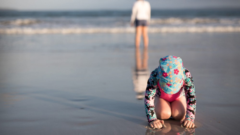 Young child playing in the sand on the beach with long sleeved swimsuit and sun safe hat.