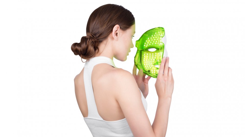 A young woman holds a facial light therapy mask up to her face.