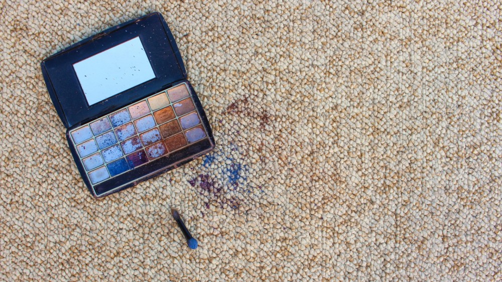 An eyeshadow palette has fallen to the floor and shades have busted over carpet.