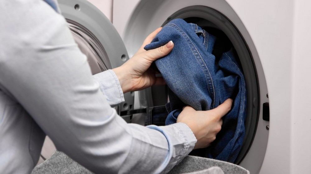 A woman in a light-colored shirt puts jeans into a front-loading washer