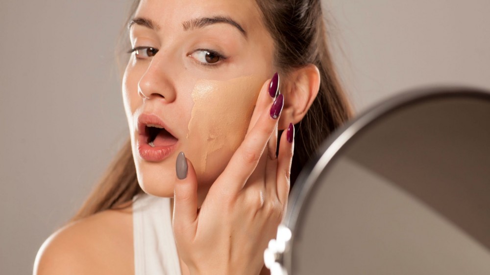 A young woman applies liquid foundation on her face with her fingers in front of a mirror.