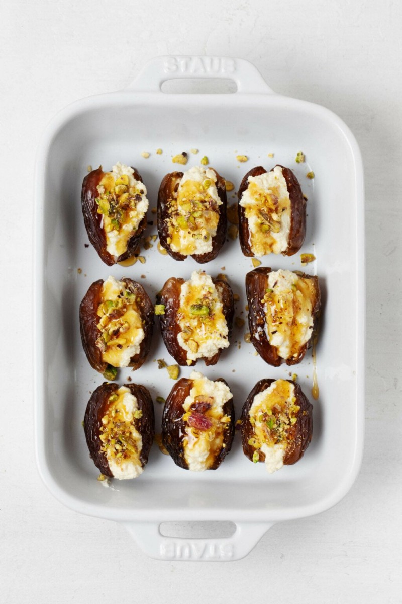 A white, rectangular porcelain container holds stuffed dates with vegan cashew cheese, chopped pistachio nuts, and a drizzle of syrup. It rests on a white surface.