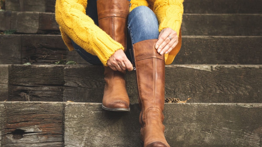 A woman puts on a pair of riding boots while sitting on a step.