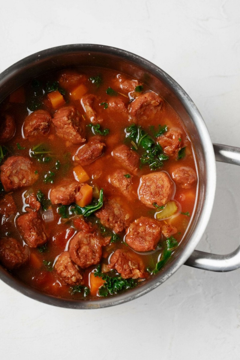 An overhead image of a soup pot, which is filled with a rustic mixture of vegetables, tomato, broth, and vegan sausage slices.