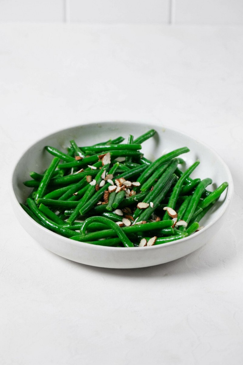 An image of a white ceramic bowl, which has been filled with brightly colored green beans and topped with sliced almonds and a vinaigrette.